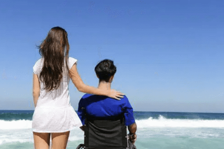 man in wheelchair with woman standing next to him in front of the ocean